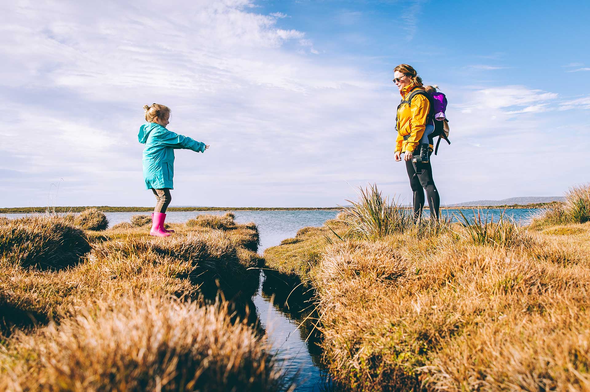 woman in yellow jacket standing across small river from girl wearing blue hacket